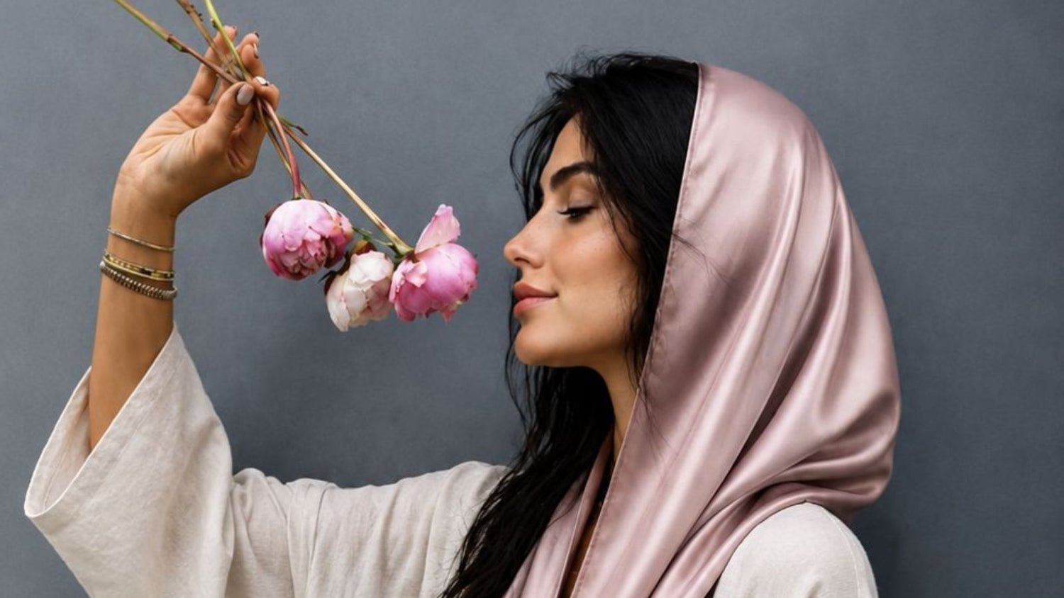 A woman wearing a pink mulberry silk hijab holding pink flowers against a gray background