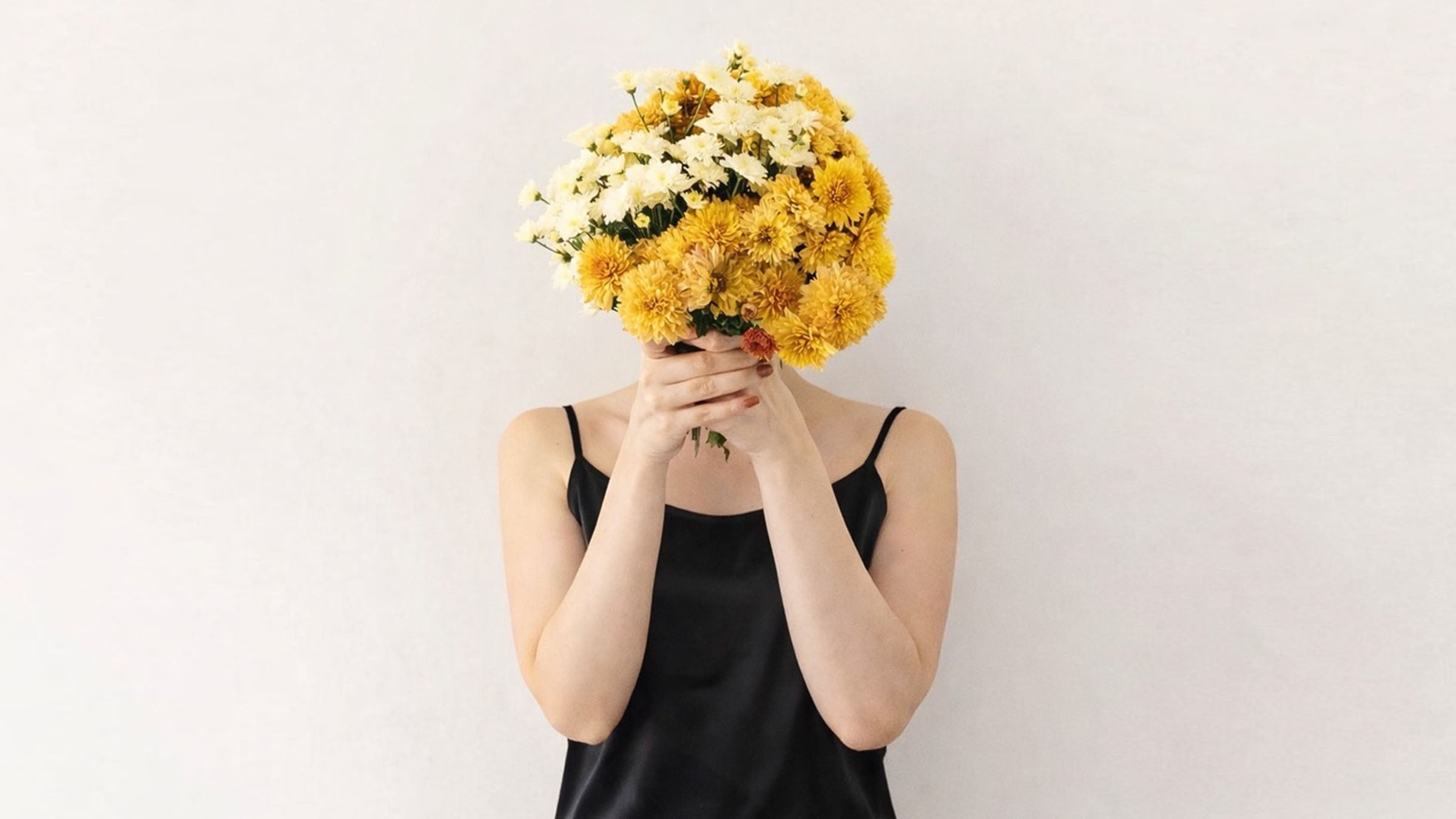 Woman wearing mulberry silk black camisole and holding a bouquet of yellow and white flowers in front of their face on a plain white background.