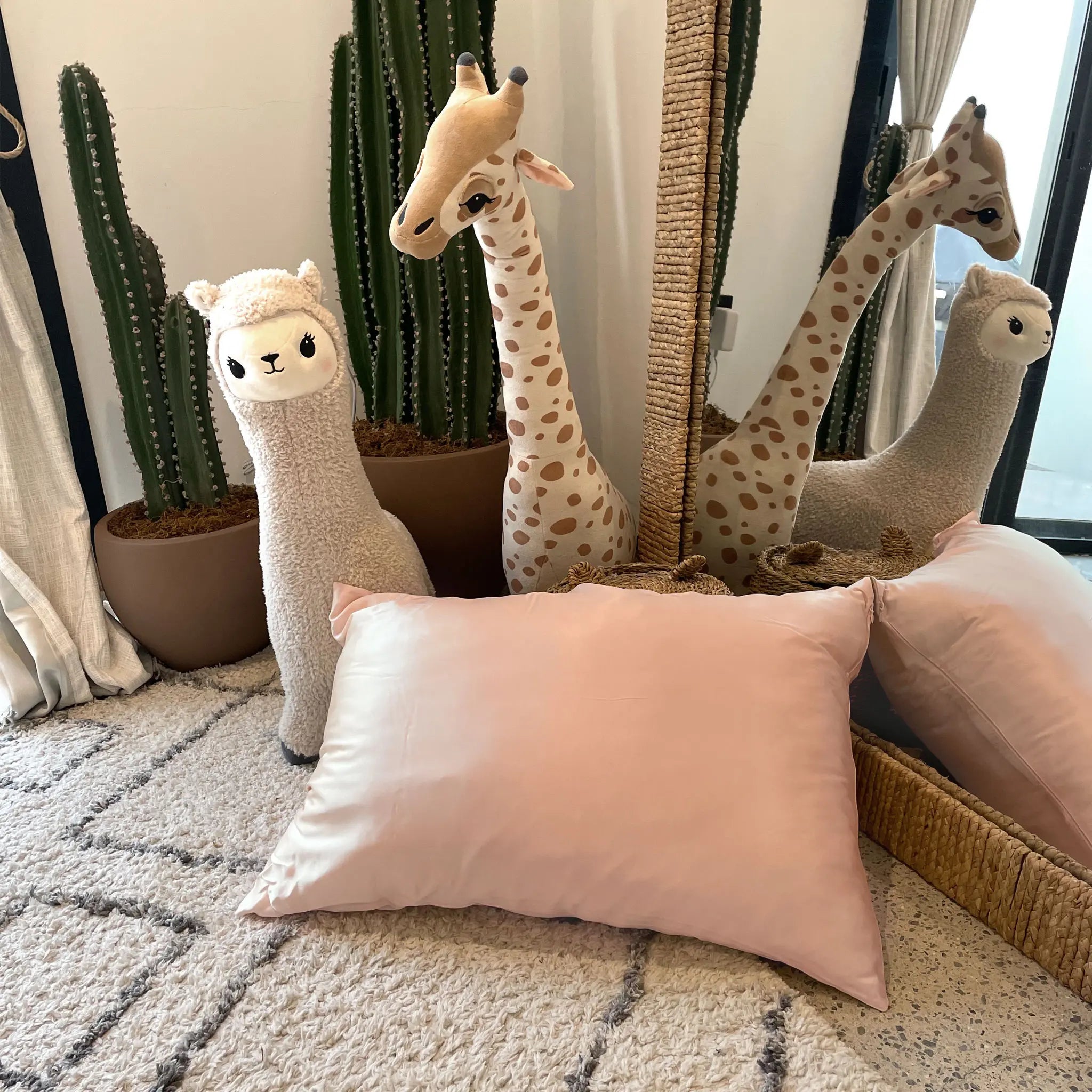 Decorative pillow in Soft Blush silk pillowcase and plush toys including a llama and giraffe on a textured surface with plants in the background. — Silk Souq pure mulberry silk home decor.