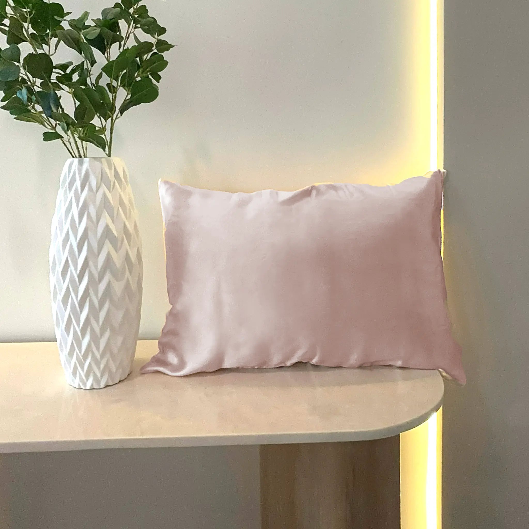 Pillow with Soft Blush silk pillowcase on a marble surface with a white vase containing greenery against a neutral wall. — Silk Souq pure mulberry silk home decor.