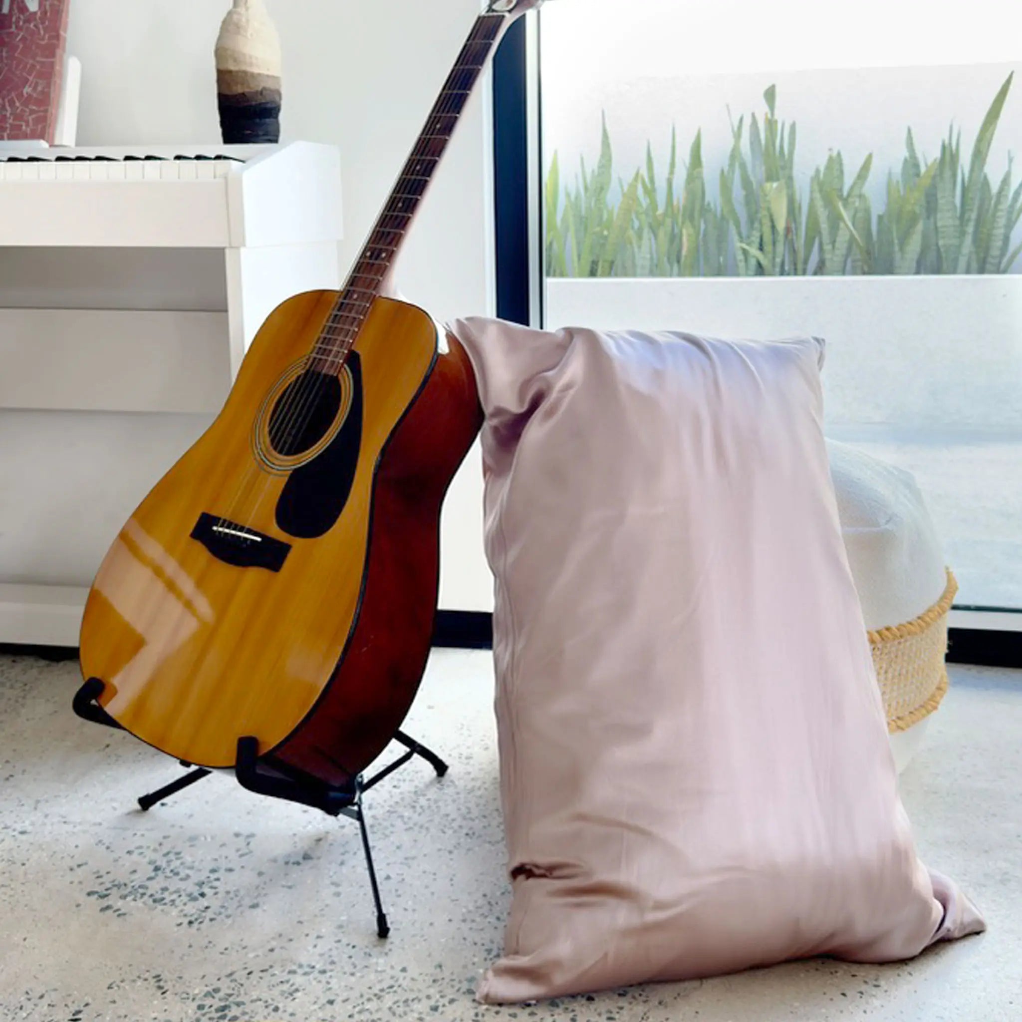 Acoustic guitar on a stand next to a pillow with Pure mulberry silk pillowcase in Taupe in a room with a window view — Silk Souq 22 momme luxury silk pillowcase