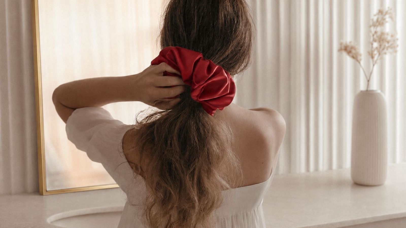 A woman with long brown hair seen from behind, wearing a large red fabric scrunchie to tie her hair, standing in a bright room with a window, curtains, and a white vase on a table.