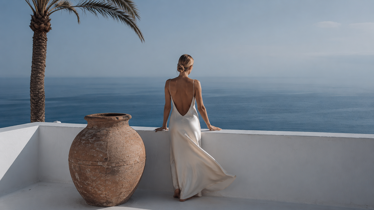 Woman in a white silk dress sitting on a ledge overlooking the ocean with a palm tree in the background.