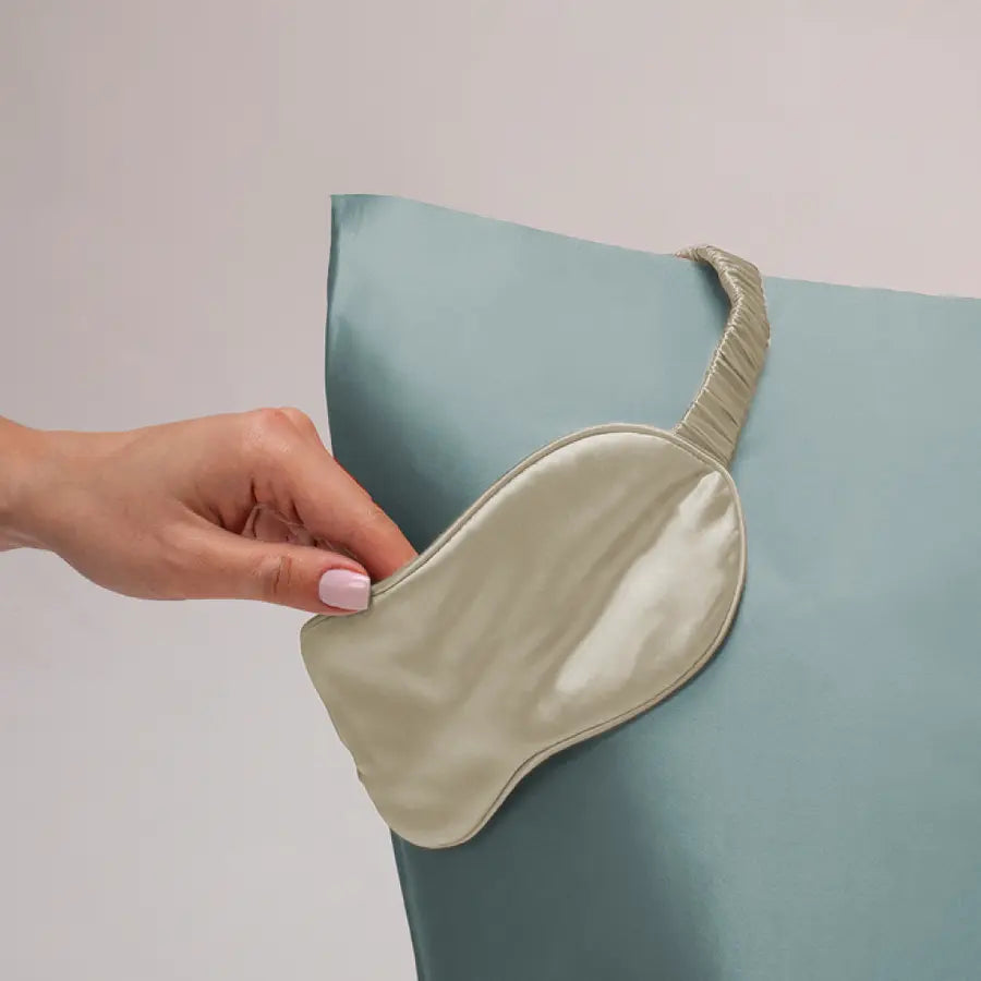 A female hand holding a pure mulberry silk sleep mask that is hanging on a pillow with pure mulberry silk pillowcase