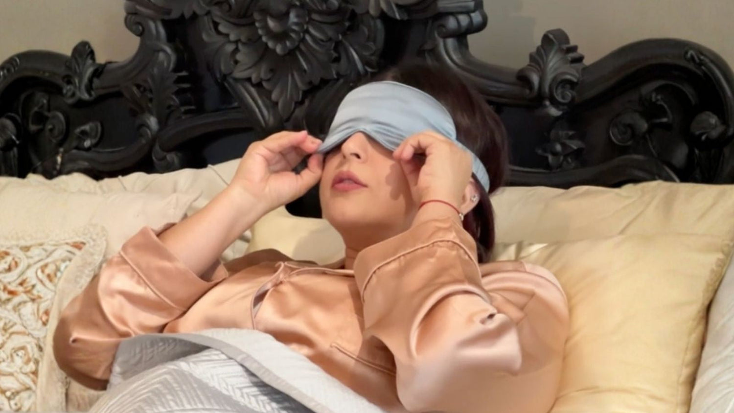 Woman wearing a silk pyjamas and silk sleep mask ilying on silk pillows on bed, looking relaxed.